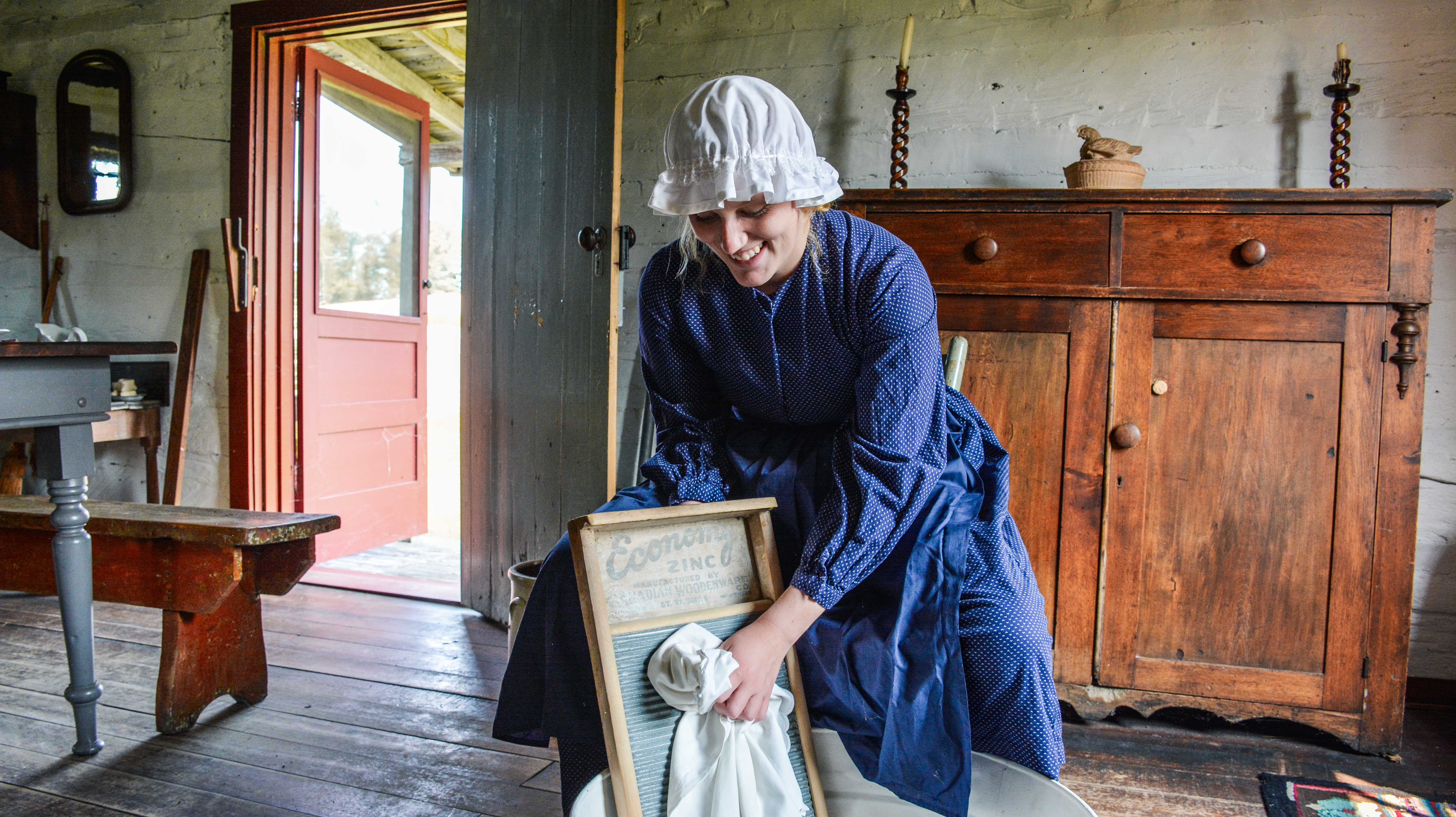 A young woman in a blud dress smiles while washing a white cloth using a washboard.