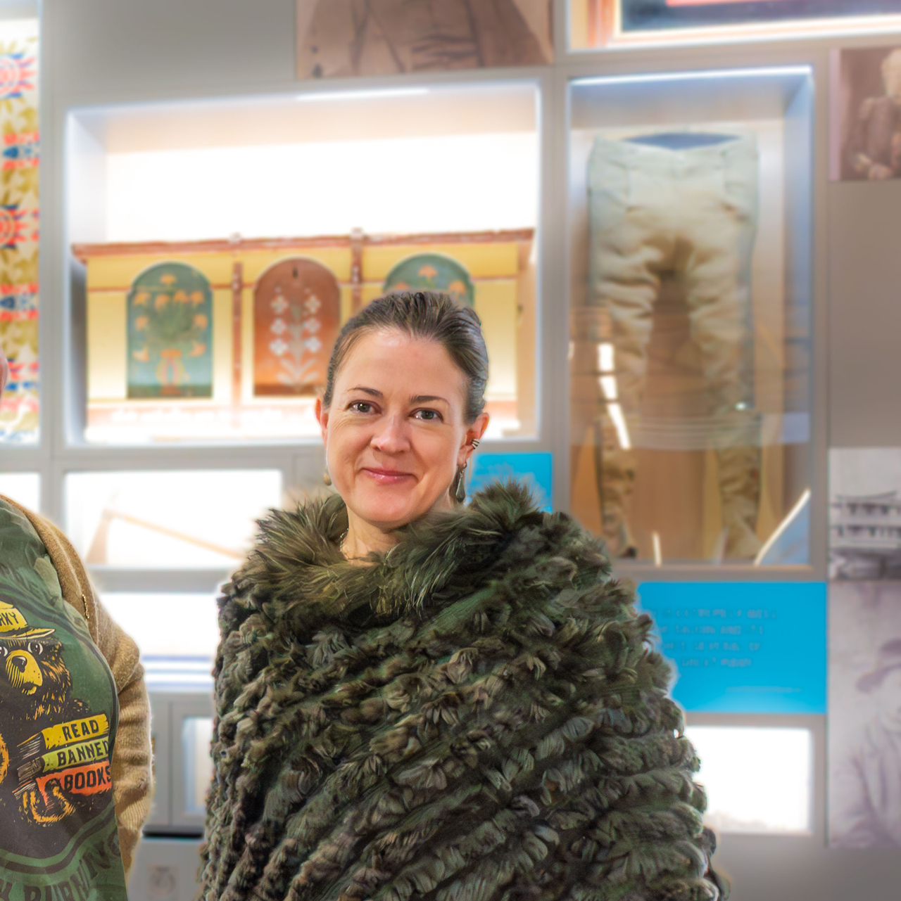 Person wearing a thick, textured fur-like shawl standing indoors in front of a display case. The background includes framed artwork, historical garments, and informational panels arranged on a wall.