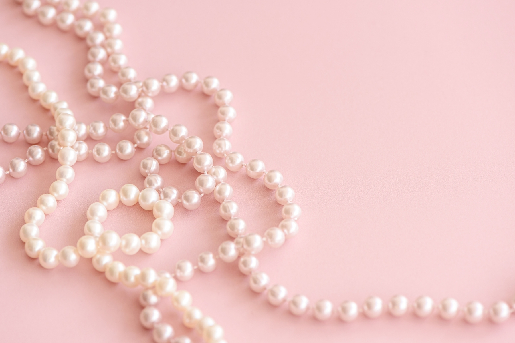 Several strands of white pearls arranged loosely on a soft pink background, creating overlapping loops and curves. The pearls have a glossy finish, reflecting light for a delicate and elegant appearance.