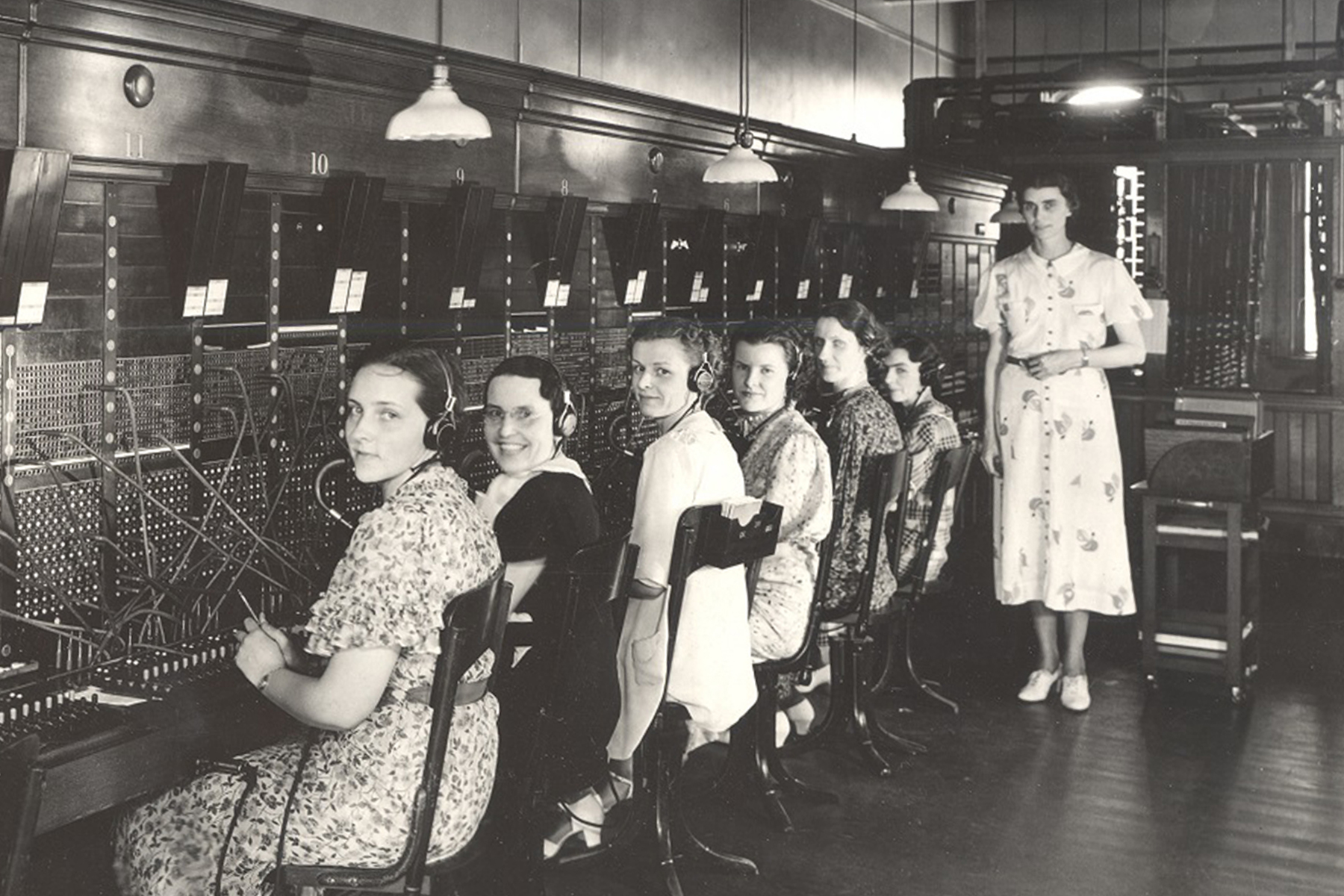 Historic photo of a telephone switchboard room with multiple operators seated in a row at wooden desks, wearing headsets and working with cables plugged into large switchboards. Overhead lamps hang above the workstations, and a person stands nearby observing.
