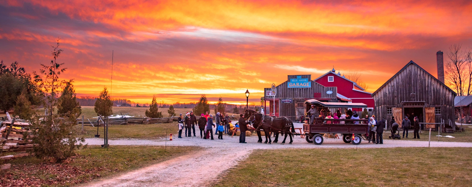 Horse-drawn wagon carrying people along a gravel path at sunset, with a red barn and wooden buildings in the background.
