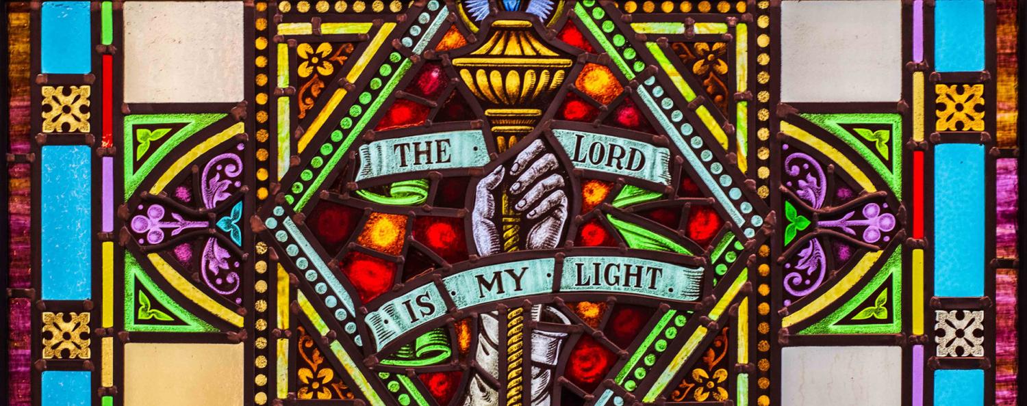 Close cropped photo of a brightly lit, stained-glass window. The window features a hand holding a torch and text stating "The Lord Is My Light".