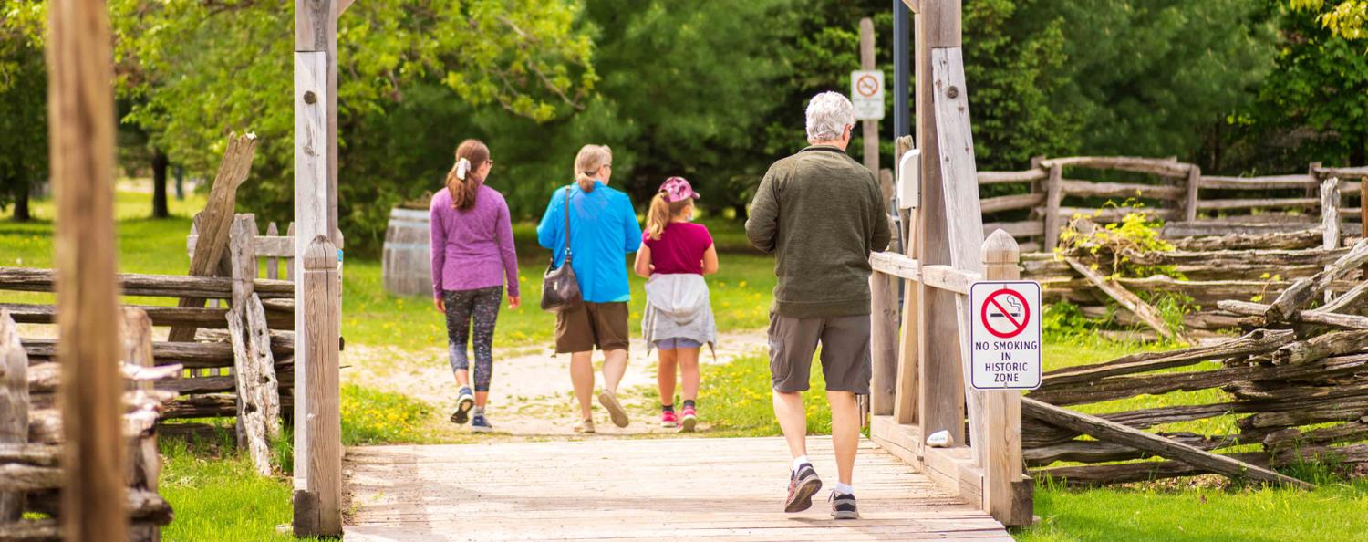 Four people—two adults and two children—walk away on a wooden walkway in a park-like setting. The walkway has rustic wooden fences on both sides, and a sign on the right reads 'NO SMOKING HISTORIC ZONE.' Green trees and grass fill the background, indicating a natural or historical area.