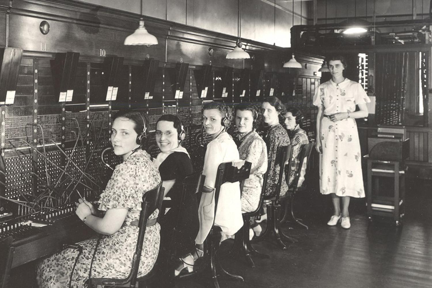 Historic photo of a telephone switchboard room with multiple operators seated in a row at wooden desks, wearing headsets and working with cables plugged into large switchboards. Overhead lamps hang above the workstations, and a person stands nearby observing.