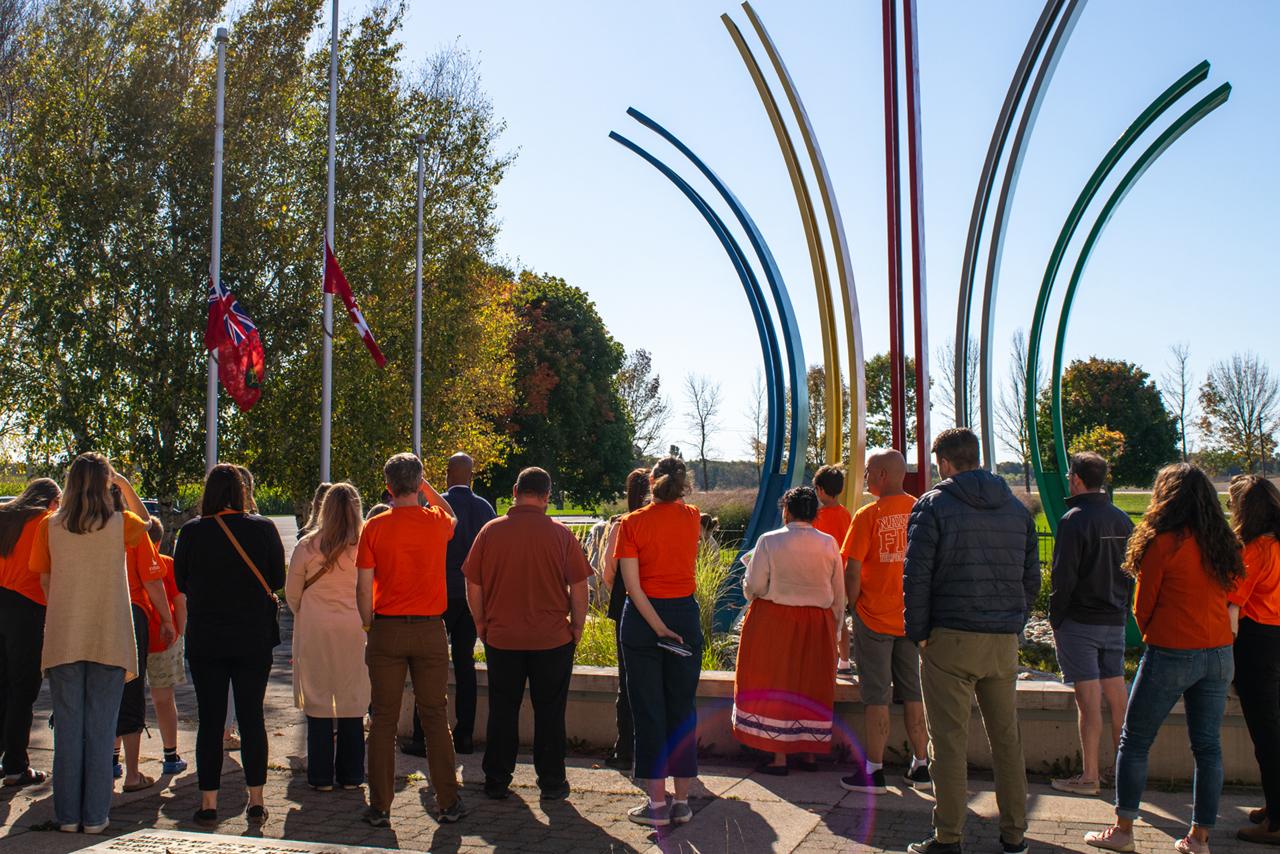 Group of people standing in a circle outdoors near a colorful metal sculpture with tall curved poles. Most individuals are wearing orange shirts, and two flags are at half-mast on flagpoles to the left. Trees with autumn foliage and a clear blue sky are in the background.