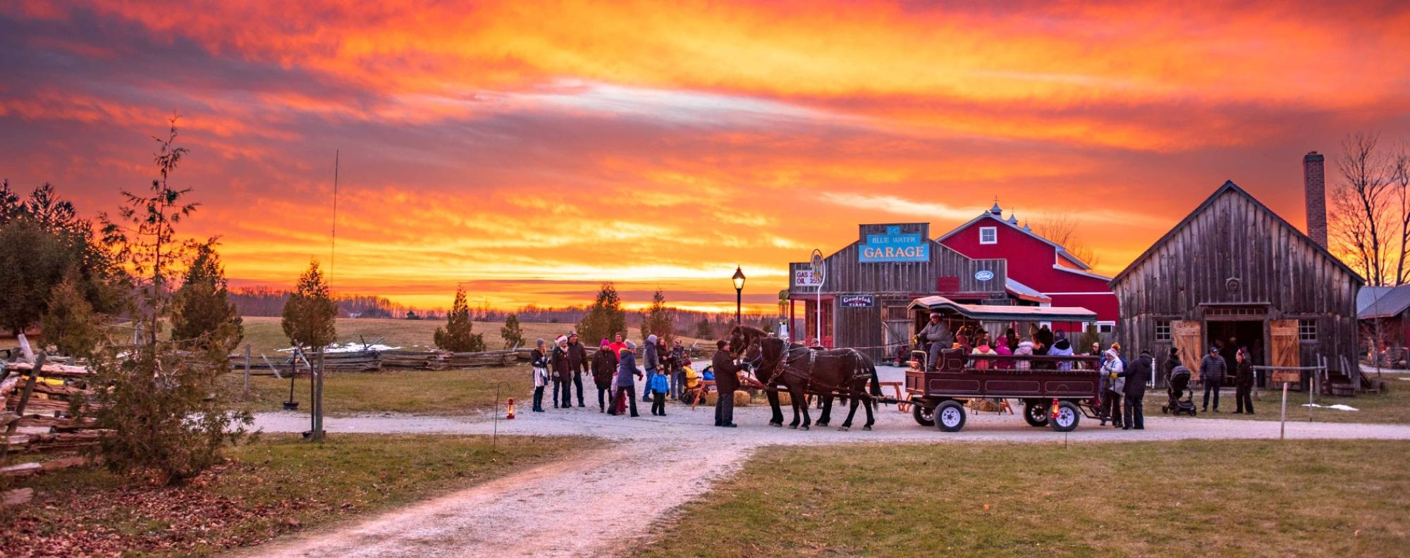 Horse-drawn wagon carrying people along a gravel path at sunset, with a red barn and wooden buildings in the background.