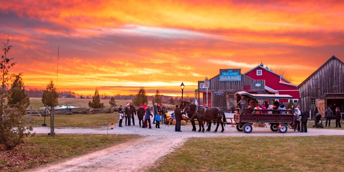 Horse-drawn wagon carrying people along a gravel path at sunset, with a red barn and wooden buildings in the background.