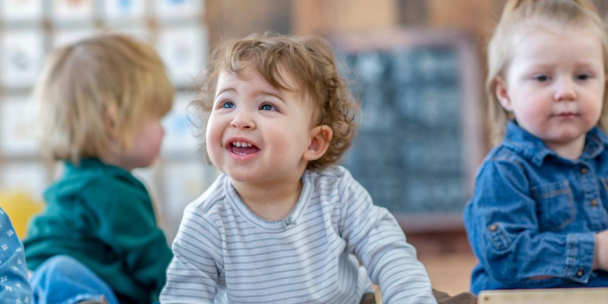 Four young children sitting indoors on the floor, wearing casual long-sleeve tops, with a wooden toy and chalkboard visible in the background.