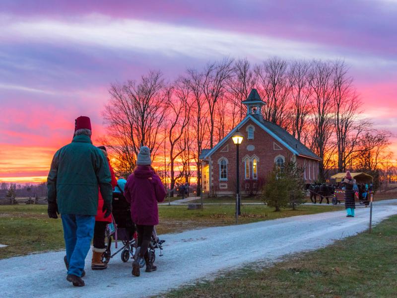 People walking along a gravel path past a small brick church at sunset, with vibrant pink and orange skies and bare trees in the background.