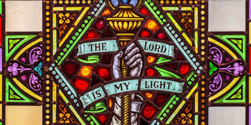 Close cropped photo of a brightly lit, stained-glass window. The window features a hand holding a torch and text stating "The Lord Is My Light".