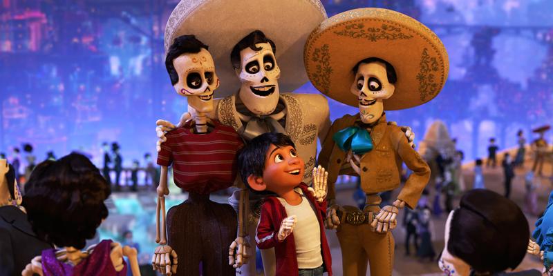 Scene from the animated movie 'Coco' featuring a young boy in a red jacket and white shirt looking up at several skeletal characters dressed in traditional Mexican attire, including sombreros and suits. The background is filled with festive lights and blurred figures, evoking a celebratory atmosphere.