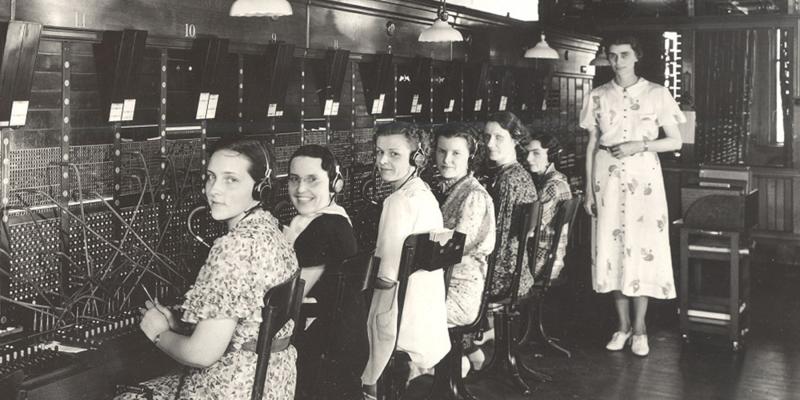 Historic photo of a telephone switchboard room with multiple operators seated in a row at wooden desks, wearing headsets and working with cables plugged into large switchboards. Overhead lamps hang above the workstations, and a person stands nearby observing.