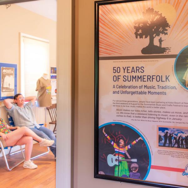 A room with two people seated on chairs to the left and a large poster on the right titled "50 YEARS OF SUMMERFOLK." The poster includes text about the festival's history and features three images: a person holding a microphone, a performer with a guitar, and a group performing on stage.