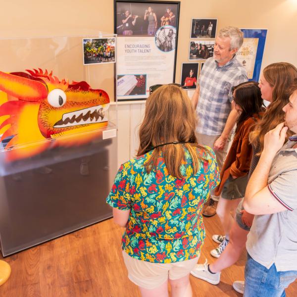 A group of people stand in a room with wooden floors, observing an exhibit featuring a colorful dragon head inside a transparent display case. The walls are decorated with posters and photographs, and a mannequin wearing a t-shirt is visible on the left side.