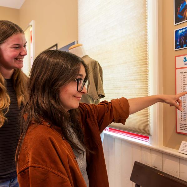 Two individuals stand in front of a wall display titled "Summerfolk Name Game." One person is pointing at the poster, which lists names under "First Name" and "Last Name" headings. Above the poster are two photographs, and a small placard is positioned below it.