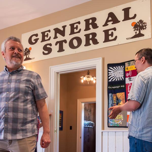 Interior of a building with a sign reading "GENERAL STORE" above a doorway. Two people are present—one on the left in a plaid shirt and beige pants, and one on the right in a striped shirt and jeans. The walls are decorated with framed pictures and colorful quilts.
