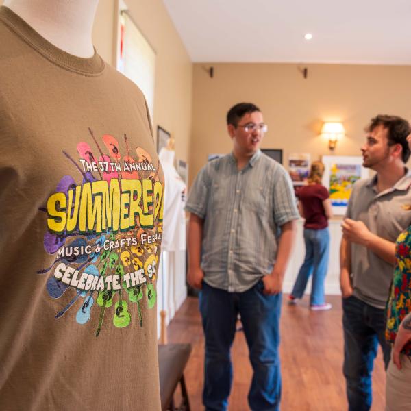 A room with several people standing and conversing on a wooden floor. In the foreground, a mannequin wears a brown t-shirt with colorful text reading: "The 37th Annual SUMMERFOLK Music & Crafts Festival". Artwork is displayed on the walls in the background.
