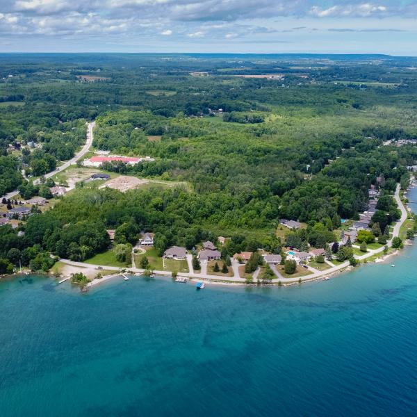 Aerial view of a shoreline community with houses along clear blue water and dense forest behind.