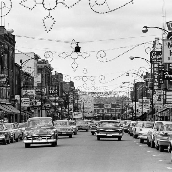Black‑and‑white photo of a busy mid‑century downtown street lined with cars, shops, and hanging decorations.