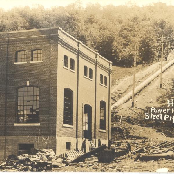 Historic photo of a brick hydro power house beside a long steel pipeline running up a wooded hillside.