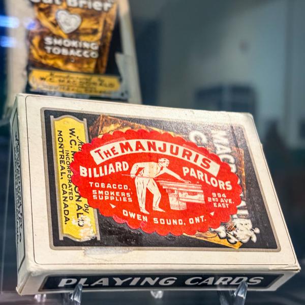 Vintage playing-card box labeled ‘The Manjuris Billiard Parlors’ displayed in a museum case.